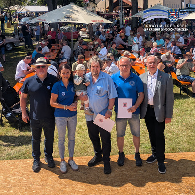 Gruppenbild bei einer Freiluftveranstaltung mit vielen Besuchern an Bierbänken im Hintergrund. Im Vordergrund stehen (v. li.) Jürgen Liebermann (Bürgermeister Schwarzenborn) mit Sonnenhut, Christin Ziegler (MdL) in blauem Poloshirt, Fred Ziegler mit Enkelkind auf dem Arm, Karsten Itzenhäuser mit Urkunde in der Hand sowie Erster Kreisbeigeordneter Jürgen Kaufmann im grauen Sakko. Die Gruppe steht auf einer Holzfläche vor den Zuschauern. Rechts oben ist das Logo des Schwalm-Eder-Kreises mit dem Schriftzug „Der Kreis mit den schönsten Ecken“ eingeblendet.