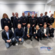 Das Foto zeigt eine Gruppe von Feuerwehrangehörigen und Vertreterinnen und Vertretern aus Verwaltung und Politik, die gemeinsam für ein Gruppenbild zusammengestanden haben. Die Personen in Uniform tragen Auszeichnungen und stehen in einer Reihe im Hintergrund, während weitere Personen in Alltags- oder Businesskleidung davor in einer Hocke- oder Kniesitzposition posieren. Alle blicken lächelnd in die Kamera. Die Szene wirkt feierlich und dokumentiert die Verleihung der Goldenen Brandschutzehrenzeichen.