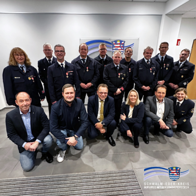Das Foto zeigt eine Gruppe von Feuerwehrangehörigen und Vertreterinnen und Vertretern aus Verwaltung und Politik, die gemeinsam für ein Gruppenbild zusammengestanden haben. Die Personen in Uniform tragen Auszeichnungen und stehen in einer Reihe im Hintergrund, während weitere Personen in Alltags- oder Businesskleidung davor in einer Hocke- oder Kniesitzposition posieren. Alle blicken lächelnd in die Kamera. Die Szene wirkt feierlich und dokumentiert die Verleihung der Goldenen Brandschutzehrenzeichen.