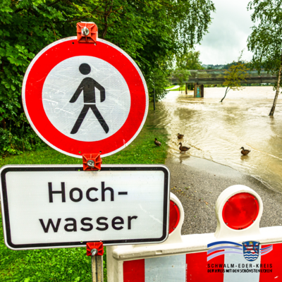 Ein rot-weißes Warnschild mit einem Fußgänger-Verbotssymbol und dem Schriftzug „Hochwasser“ steht vor einer gesperrten Straße. Die Straße ist überflutet, Enten schwimmen auf dem Wasser. Im Hintergrund sind Bäume, eine Brücke und bewölkter Himmel zu sehen. Unten rechts befindet sich das Logo des Schwalm-Eder-Kreises.