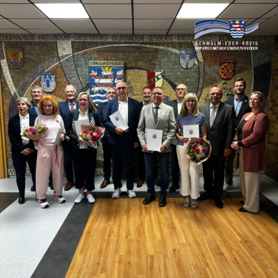 Gruppenfoto in der Kreisverwaltung des Schwalm-Eder-Kreises: Mehrere Frauen und Männer stehen vor einem Wandmosaik mit den Wappen des Schwalm-Eder-Kreises und seiner Kommunen. In der ersten Reihe halten einige der Geehrten ihre Ehrenbriefe des Landes Hessen sowie Blumensträuße in den Händen. Gemeinsam mit Landrat Winfried Becker und weiteren Gästen wurden sie für ihr langjähriges ehrenamtliches Engagement ausgezeichnet. Oben rechts ist das Logo des Schwalm-Eder-Kreises eingeblendet.