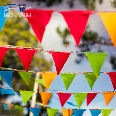 Bunte Wimpelketten in Rot, Grün, Orange und Blau hängen unter freiem Himmel und flattern im Wind. Im Hintergrund sind verschwommen Bäume und der Himmel zu erkennen. Links oben ist das Logo des Schwalm-Eder-Kreises mit dem Schriftzug „Der Kreis mit den schönsten Ecken“ eingeblendet.