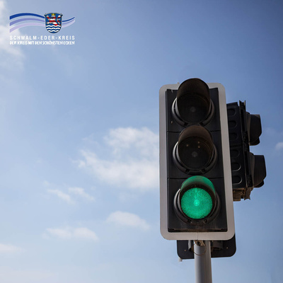 Nahaufnahme einer Ampel vor blauem Himmel mit leichten Wolken. Das grüne Licht ist eingeschaltet. Links oben ist das Logo des Schwalm-Eder-Kreises mit dem Schriftzug „Der Kreis mit den schönsten Ecken“ eingeblendet.