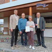 Vier Erwachsene stehen vor einem Wohnhaus mit überdachter Terrasse und blicken in die Kamera. Eine Frau in der Mitte hält eine Urkunde in den Händen. Zwei Männer tragen Anzug und Krawatte, die beiden anderen Personen sind in Alltagskleidung gekleidet. Oben rechts ist das Logo des Schwalm-Eder-Kreises eingeblendet.
