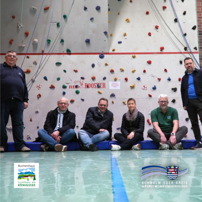 Sechs Erwachsene befinden sich in einer Sporthalle vor einer Kletterwand mit bunten Griffen. Vier Personen sitzen nebeneinander auf einer blauen Matte am Boden, zwei weitere stehen seitlich daneben. Auf der Wand ist der Schriftzug „Red Rooster“ zu sehen. Unten im Bild sind die Logos von Buchenhaus Schönau am Königssee und dem Schwalm-Eder-Kreis eingeblendet.
