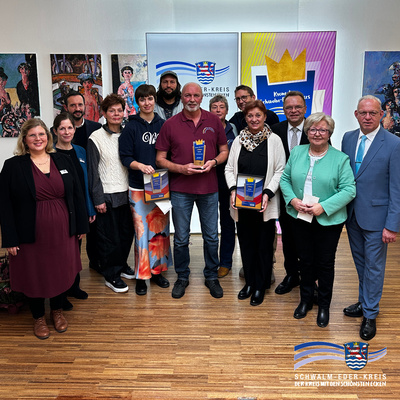 Auf dem Foto steht eine Gruppe von etwa dreizehn Personen in einem hellen Raum vor einer Wand mit farbenfrohen Gemälden und Bannern des Schwalm-Eder-Kreises. Drei Personen in der Mitte halten Trophäen in den Händen. Alle lächeln in die Kamera, und es herrscht eine feierliche, freundliche Stimmung. Im Vordergrund ist der Holzboden zu sehen, im Hintergrund das Logo des Schwalm-Eder-Kreises mit dem Schriftzug „Der Kreis mit den schönsten Ecken“.