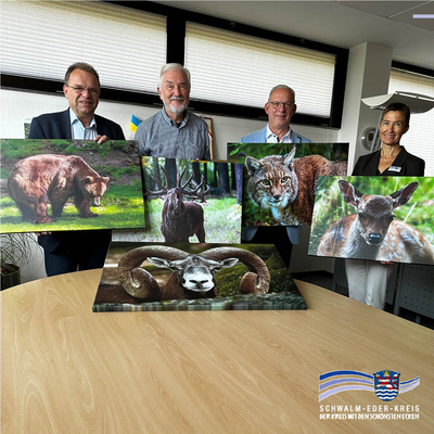 Von links nach rechts: Landrat Winfried Becker hält ein Foto von einem Bären, daneben steht Gerhard Schmoll mit einem Bild von einem Hirsch mit Geweih. Der Erste Kreisbeigeordnete Jürgen Kaufmann zeigt ein Foto von einem Luchs. Rechts steht Kerstin Schwab, Leiterin des Eigenbetriebs, mit einem Bild von einem Hirschkalb. Auf dem Tisch vor ihnen liegt zusätzlich ein Foto von einem Mufflon mit großen Hörnern. Rechts unten ist das Logo des Schwalm-Eder-Kreises mit der Aufschrift „Schwalm-Eder-Kreis – Der Kreis mit den schönsten Ecken“.