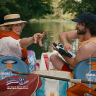 Auf dem Bild sitzen eine ältere Frau und ein jüngerer Mann gemeinsam in einem Tretboot auf einem ruhigen, grünen Fluss. Beide tragen Rettungswesten. Die Frau mit Strohhut zeigt mit ausgestrecktem Arm in eine Richtung, während der Mann mit Bart und Kappe sie anschaut und ein kleines technisches Gerät in den Händen hält. Hinter ihnen umgeben dicht bewachsene Bäume das Wasser. Auf dem Bild sitzen eine ältere Frau und ein jüngerer Mann gemeinsam in einem Tretboot auf einem ruhigen, grünen Fluss. Beide tragen Rettungswesten. Die Frau mit Strohhut zeigt mit ausgestrecktem Arm in eine Richtung, während der Mann mit Bart und Kappe sie anschaut und ein kleines technisches Gerät in den Händen hält. Hinter ihnen umgeben dicht bewachsene Bäume das Wasser.