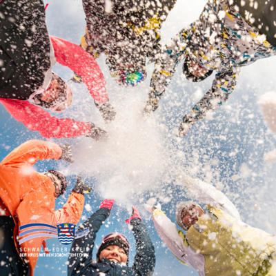 Eine Gruppe von Menschen in bunter Winterkleidung steht im Kreis und wirft lachend Schnee in die Luft. Der Blickwinkel kommt von unten, gegen den blauen Himmel, sodass die ausgestreckten Arme und der fliegende Schnee im Mittelpunkt stehen. Unten links ist das Logo des Schwalm-Eder-Kreises zu sehen.