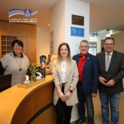 Vier Erwachsene stehen an der Rezeption eines Hotels und blicken in die Kamera. Eine Frau steht hinter dem Empfangstresen, auf dem sich eine kleine dekorative Osterdekoration mit Pflanzen und Figuren befindet. Drei weitere Personen stehen davor nebeneinander. Im Hintergrund sind ein Schlüsselschrank, ein Flur sowie an der Wand angebrachte Auszeichnungen zu sehen. Oben links befindet sich das Logo des Schwalm-Eder-Kreises mit dem Schriftzug „Der Kreis mit den schönsten Ecken“.