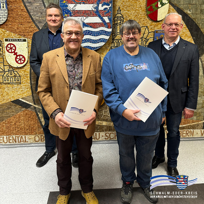 Vor einem großen Wandmosaik mit den Wappen des Schwalm-Eder-Kreises und seiner Kommunen stehen vier Männer und blicken lächelnd in die Kamera. In der ersten Reihe halten Holger Raude (links) und Jörg Vollbrecht (rechts) jeweils eine weiße Mappe mit dem Logo des Schwalm-Eder-Kreises. Dahinter stehen Lars Werner (links) und Jürgen Kaufmann (rechts), beide in dunklen Sakkos. Unten rechts im Bild ist das Logo des Schwalm-Eder-Kreises mit dem Schriftzug „Der Kreis mit den schönsten Ecken“ eingeblendet.