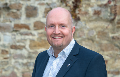 Porträtfoto von Jan Rauschenberg, Kreistagsmitglied im Schwalm-Eder-Kreis. Er steht lächelnd vor einer Natursteinmauer und trägt ein dunkles Sakko über einem karierten Hemd. Am Revers seines Sakkos ist eine kleine Anstecknadel zu sehen. Die Aufnahme wirkt freundlich und professionell.