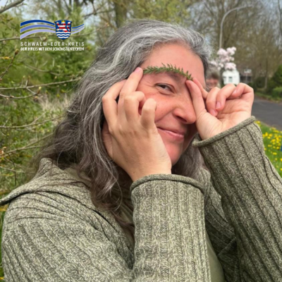 Eine Frau mit langen grauen Haaren steht im Freien vor Bäumen und lächelt in die Kamera. Mit beiden Händen hält sie einen kleinen grünen Zweig über ihre Augenbrauen. Im Hintergrund sind Frühlingsblüten zu sehen, oben links ist das Logo des Schwalm-Eder-Kreises eingeblendet.