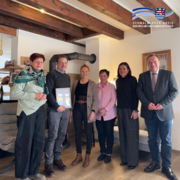 In einem gemütlich eingerichteten Raum mit sichtbaren Holzbalken an der Decke stehen sechs Erwachsene nebeneinander und blicken in die Kamera. Ein Mann in der Mitte hält eine gerahmte Urkunde in den Händen. Die übrigen Personen stehen links und rechts neben ihm. Im Hintergrund sind eine Sitzecke, ein Ofenrohr sowie dekorative Elemente an den Wänden zu sehen. Oben rechts im Bild befindet sich das Logo des Schwalm-Eder-Kreises mit dem Schriftzug „Der Kreis mit den schönsten Ecken“.