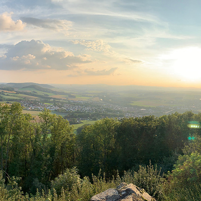 Panoramablick vom Heiligenberg: Im Vordergrund eine alte Steinmauer mit mehreren Holzbänken und -tischen auf einer grünen Wiese, umgeben von Bäumen. Dahinter breitet sich eine weite Landschaft mit Feldern, Wäldern und kleinen Ortschaften aus. Am Horizont geht die Sonne unter, taucht den Himmel in warme Gelb- und Orangetöne und wirft weiches Licht über die Szenerie. Links und rechts rahmen Bäume den Blick ein.
