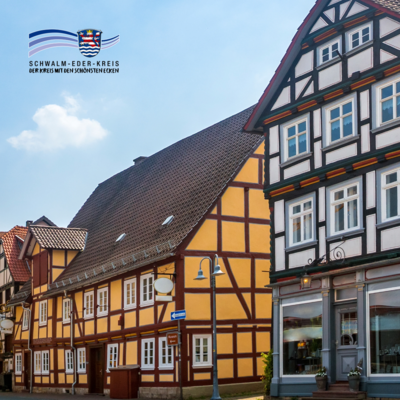 Zu sehen ist eine Straße mit historischen Fachwerkhäusern in warmen Gelb- und Brauntönen. Die Gebäude haben weiße Fensterrahmen und steile Ziegeldächer, eines zeigt schwarz-weißes Fachwerk mit farbigen Akzenten. Im Vordergrund steht eine Straßenlaterne, der Himmel ist blau mit wenigen Wolken. Oben links ist das Logo des Schwalm-Eder-Kreises eingeblendet.