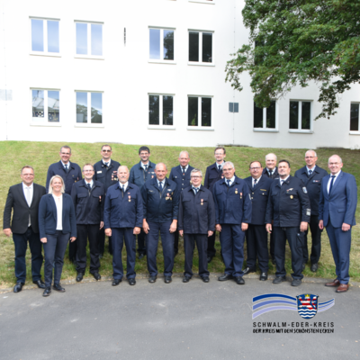 Gruppenfoto vor einem Verwaltungsgebäude: Mehrere Feuerwehrkameraden in Uniform stehen gemeinsam mit Vertreterinnen und Vertretern aus Politik und Verwaltung auf einer Grünfläche vor einer hellen Gebäudefassade. Einige der Feuerwehrangehörigen tragen Auszeichnungen an ihren Uniformen. Das Foto entstand im Rahmen der Verleihung der Goldenen Brandschutzehrenzeichen für 40 und 50 Jahre aktiven Feuerwehrdienst. Unten rechts ist das Logo des Schwalm-Eder-Kreises eingeblendet.