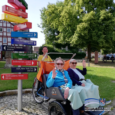 Barbara Stübing fährt eine E-Rikscha, in der Anita Lange und Käte Schönwalder sitzen. Alle drei lächeln und winken in die Kamera. Im Hintergrund steht ein bunter Wegweiser mit internationalen Städtenamen und Entfernungsangaben, daneben sind Bäume und Fachwerkhäuser zu sehen. Unten rechts ist das Logo des Schwalm-Eder-Kreises mit dem Schriftzug „Der Kreis mit den schönsten Ecken“ eingeblendet.
