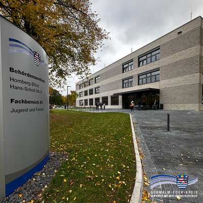 Das Bild zeigt das moderne Verwaltungsgebäude des Fachbereichs 51 – Jugend und Familie – im Behördenzentrum Homberg (Efze), Hans-Scholl-Straße 8. Links im Vordergrund steht eine große graue Hinweistafel mit dem Logo des Schwalm-Eder-Kreises und der Aufschrift: „Behördenzentrum Homberg (Efze), Hans-Scholl-Str. 8, Fachbereich 51, Jugend und Familie“. Rechts davon ist das helle, dreigeschossige Gebäude mit großen Fenstern zu sehen. Einige Personen befinden sich auf dem Vorplatz. Der Himmel ist bewölkt, auf dem Boden liegen herabgefallene gelbe Blätter. Unten rechts ist erneut das Logo des Schwalm-Eder-Kreises mit dem Slogan „Der Kreis mit den schönsten Ecken“ abgebildet.