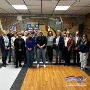 „Gruppenfoto von 16 Erwachsenen, die nebeneinander in einem Innenraum stehen. Sie blicken in die Kamera. Im Hintergrund befindet sich eine große Wandgestaltung mit Wappen und Ortsnamen des Schwalm-Eder-Kreises. Der Boden ist teilweise aus Holz, teilweise aus hellem Belag. Rechts unten ist das Logo des Schwalm-Eder-Kreises zu sehen.“