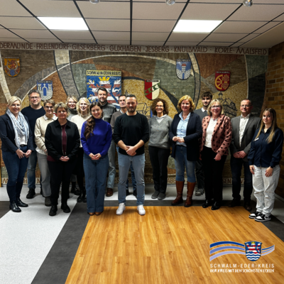 „Gruppenfoto von 16 Erwachsenen, die nebeneinander in einem Innenraum stehen. Sie blicken in die Kamera. Im Hintergrund befindet sich eine große Wandgestaltung mit Wappen und Ortsnamen des Schwalm-Eder-Kreises. Der Boden ist teilweise aus Holz, teilweise aus hellem Belag. Rechts unten ist das Logo des Schwalm-Eder-Kreises zu sehen.“