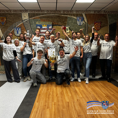 Gruppenfoto eines jubelnden Teams in einem Gebäude mit Mosaikwand im Hintergrund, auf der der Schriftzug „SCHWALM-EDER-KREIS“ sowie verschiedene Ortsnamen und Wappen zu sehen sind. Die Personen tragen weiße T-Shirts mit der Aufschrift „Hessen Meister“ und zeigen Pokale und Urkunden. Zwei Männer knien vorne in der Mitte, einer hält einen großen goldenen Siegerpokal. Unten rechts ist das Logo des Schwalm-Eder-Kreises mit dem Slogan „Der Kreis mit den schönsten Ecken“ zu sehen. Die Szene strahlt Freude, Zusammenhalt und sportlichen Erfolg aus.