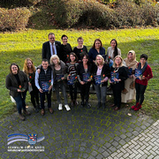 Das Bild zeigt eine Gruppe von 15 Personen, die im Freien auf einem gepflasterten Platz stehen und in die Kamera lächeln. Einige von ihnen halten Urkunden und rote Rosen in den Händen. Im Hintergrund ist eine Grünfläche mit Büschen zu sehen. Es herrscht eine fröhliche und feierliche Stimmung, die auf eine Auszeichnung oder den Abschluss einer Qualifikation hinweist. Unten links im Bild befindet sich das Logo des Schwalm-Eder-Kreises mit dem Schriftzug „Der Kreis mit den schönsten Ecken“.