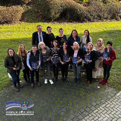 Das Bild zeigt eine Gruppe von 15 Personen, die im Freien auf einem gepflasterten Platz stehen und in die Kamera lächeln. Einige von ihnen halten Urkunden und rote Rosen in den Händen. Im Hintergrund ist eine Grünfläche mit Büschen zu sehen. Es herrscht eine fröhliche und feierliche Stimmung, die auf eine Auszeichnung oder den Abschluss einer Qualifikation hinweist. Unten links im Bild befindet sich das Logo des Schwalm-Eder-Kreises mit dem Schriftzug „Der Kreis mit den schönsten Ecken“.
