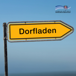 Zu sehen ist ein gelbes, pfeilförmiges Straßenschild mit schwarzem Rand und der Aufschrift „Dorfladen“. Der Pfeil zeigt nach rechts und ist an einem Metallmast befestigt. Im Hintergrund ist ein blauer Himmel zu erkennen. Oben rechts ist das Logo des Schwalm-Eder-Kreises eingeblendet.