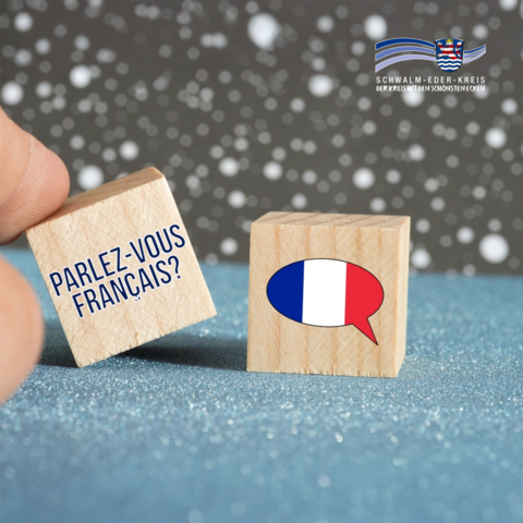 Zwei kleine Holzwürfel stehen auf einer Oberfläche. Auf einem Würfel steht der französische Satz „Parlez-vous français?“, auf dem anderen ist eine Sprechblase in den Farben der französischen Flagge abgebildet. Oben rechts ist das Logo des Schwalm-Eder-Kreises eingeblendet.