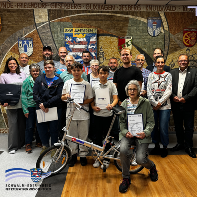 Gruppenfoto der Siegerinnen und Sieger des Stadtradelns 2025 im Schwalm-Eder-Kreis. In der vorderen Reihe stehen und sitzen mehrere Teilnehmende mit Urkunden, ein Junge hält ein silbernes Klapprad. Neben ihm eine Frau mit grauen Haaren und Brille, die ebenfalls eine Urkunde präsentiert. Hinter ihnen stehen weitere Teilnehmerinnen und Teilnehmer in mehreren Reihen. Ganz rechts außen steht Landrat Winfried Becker im dunklen Anzug, ganz links außen Tatjana Grau-Becker, Leiterin der Wirtschaftsförderung, im lila Pullover. Im Hintergrund ist ein Wandmosaik mit Wappen der Kommunen des Schwalm-Eder-Kreises zu sehen.