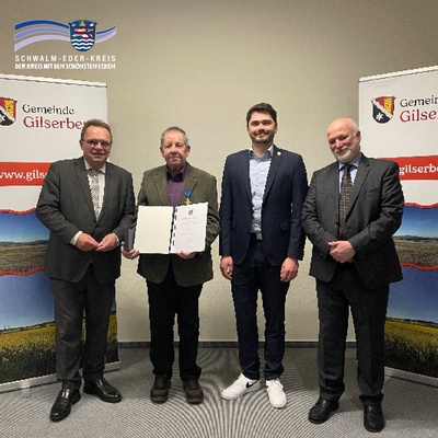 Vier Männer stehen nebeneinander vor zwei Roll-Up-Bannern mit dem Wappen und Schriftzug der Gemeinde Gilserberg. In der Mitte hält der zweite Mann von links, Klaus Böse, eine geöffnete Urkunde in den Händen und trägt eine Auszeichnung am Revers. Von links nach rechts sind zu sehen: Landrat Winfried Becker, der Geehrte Klaus Böse, Bürgermeister Lukas Daum und Philipp Zmyi-Köbel, Direktor des Amtsgerichts Schwalmstadt. Links oben im Bild befindet sich das Logo des Schwalm-Eder-Kreises mit dem Slogan „Der Kreis mit den schönsten Ecken“.