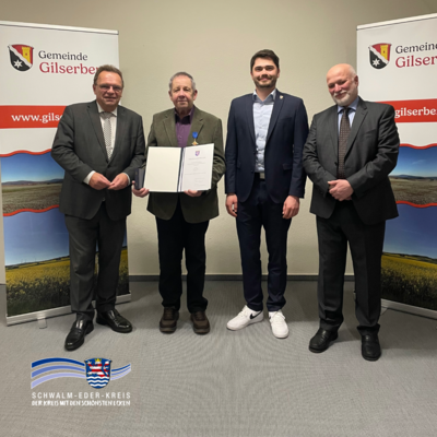 Vier Männer stehen nebeneinander vor zwei Roll-Up-Bannern mit dem Wappen und Schriftzug der Gemeinde Gilserberg. In der Mitte hält der zweite Mann von links, Klaus Böse, eine geöffnete Urkunde in den Händen und trägt eine Auszeichnung am Revers. Von links nach rechts sind zu sehen: Landrat Winfried Becker, der Geehrte Klaus Böse, Bürgermeister Lukas Daum und Philipp Zmyi-Köbel, Direktor des Amtsgerichts Schwalmstadt. Links oben im Bild befindet sich das Logo des Schwalm-Eder-Kreises mit dem Slogan „Der Kreis mit den schönsten Ecken“.
