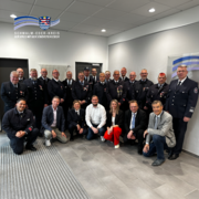 Barrierefreie Bildunterschrift: Gruppenfoto in einem hellen Innenraum: Mehrere Feuerwehrangehörige in Uniform sowie Vertreter aus Politik und Verwaltung stehen und knien gemeinsam für ein Foto. Einige der Feuerwehrleute tragen Auszeichnungen an ihren Uniformen. In der vorderen Reihe befinden sich mehrere Personen in Zivilkleidung, darunter auch eine Frau in roter Hose. Alle blicken in die Kamera. Oben links ist das Logo des Schwalm-Eder-Kreises eingeblendet.