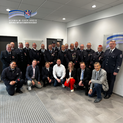 Barrierefreie Bildunterschrift: Gruppenfoto in einem hellen Innenraum: Mehrere Feuerwehrangehörige in Uniform sowie Vertreter aus Politik und Verwaltung stehen und knien gemeinsam für ein Foto. Einige der Feuerwehrleute tragen Auszeichnungen an ihren Uniformen. In der vorderen Reihe befinden sich mehrere Personen in Zivilkleidung, darunter auch eine Frau in roter Hose. Alle blicken in die Kamera. Oben links ist das Logo des Schwalm-Eder-Kreises eingeblendet.