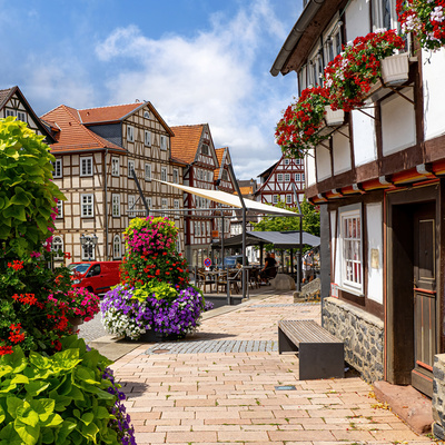 Blick auf den historischen Marktplatz in Homberg (Efze) an einem sonnigen Tag. Links und im Hintergrund sind prächtige Fachwerkhäuser mit roten Ziegeldächern zu sehen. Vor den Gebäuden blühen bunte Blumen in großen Kübeln, darunter rote, pinke, violette und weiße Blüten. Rechts im Vordergrund ein Fachwerkhaus mit Blumenkästen voller roter Geranien an den Fenstern. In der Mitte des Platzes stehen Sitzgelegenheiten unter Sonnensegeln und Sonnenschirmen. Eine entspannte, sommerliche Atmosphäre prägt die Szene.