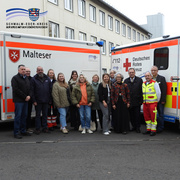 Eine Gruppe von 15 Personen steht lächelnd zwischen zwei Rettungswagen des Malteser Hilfsdienstes und des Deutschen Roten Kreuzes. Zu sehen sind Mitarbeitende beider Organisationen sowie Vertreterinnen und Vertreter der Kreisverwaltung Schwalm-Eder. Die Szene spielt sich auf einem Parkplatz vor einem Verwaltungsgebäude ab. Im Bildhintergrund das mehrstöckige Gebäude mit weißen Fassaden und vielen Fenstern. Oben links ist das Logo des Schwalm-Eder-Kreises mit dem Schriftzug „Der Kreis mit den schönsten Ecken“ eingeblendet.