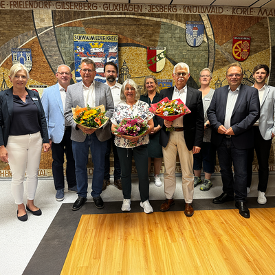Gruppenfoto in einem Gebäude der Kreisverwaltung Schwalm-Eder vor einer Wand mit Wappen und Schriftzug „Schwalm-Eder-Kreis“. Anlass ist die Ehrung von Kolleginnen und Kollegen. Zehn Personen stehen nebeneinander, drei von ihnen halten Blumensträuße. Abgebildet sind (v. li.): Stefanie Fuchs (Fachbereichsleitung Grundstücks- und Gebäudewirtschaft), Erster Kreisbeigeordneter Jürgen Kaufmann, Thomas Buck (Fachbereich Landwirtschaft und Landentwicklung) mit Blumenstrauß, Ingvar Grebe (Fachbereichsleitung Landwirtschaft und Landentwicklung), Birgit Peppler (Fachbereich Grundstücks- und Gebäudewirtschaft) mit Blumenstrauß, Ninette Bender (Fachbereich Grundstücks- und Gebäudewirtschaft), Dirk Siebert (Fachbereichsleitung Digitalisierung), Angelika Roth (Personalrat), Landrat Winfried Becker sowie Personalchef Tobias Heipel. Alle lächeln in die Kamera.