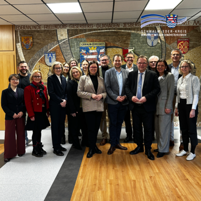 Mehrere Erwachsene stehen in einem Innenraum nebeneinander und blicken in die Kamera. Sie tragen überwiegend Business- oder gepflegte Alltagskleidung. Im Hintergrund ist eine große Wandgestaltung mit dem Schriftzug „Schwalm-Eder-Kreis“ sowie Wappen verschiedener Städte und Gemeinden zu sehen. Oben rechts ist zusätzlich das Logo des Schwalm-Eder-Kreises eingeblendet.